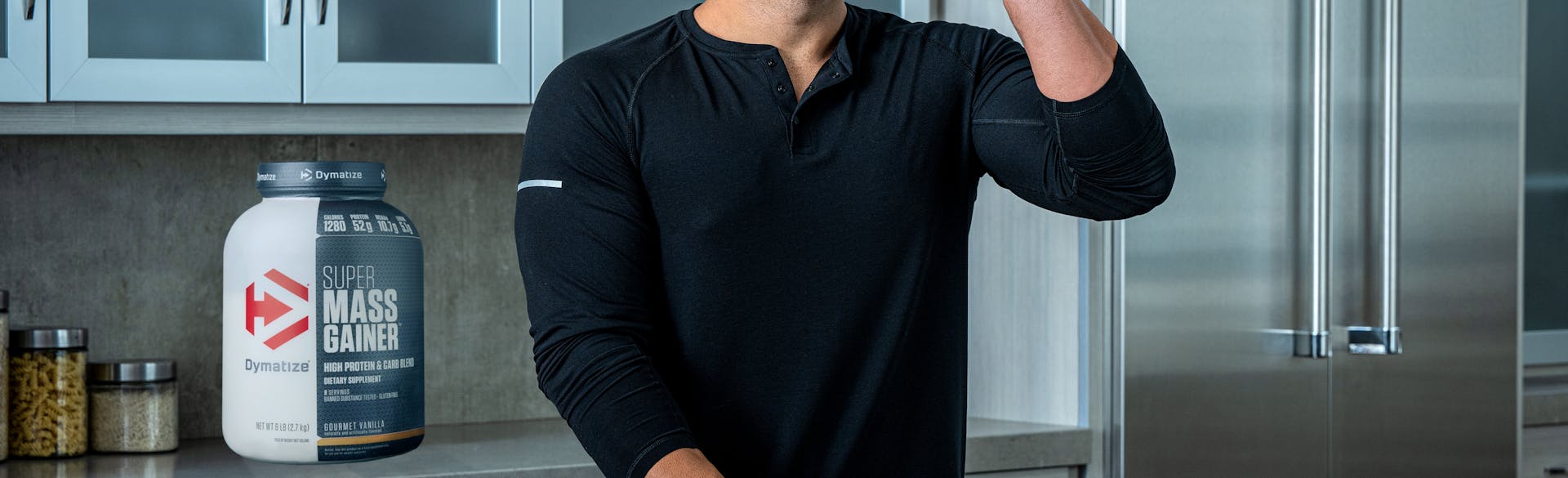 A man in a kitchen, he is drinking a Dymatize Super Mass Gainer shake out of a shaker bottle.