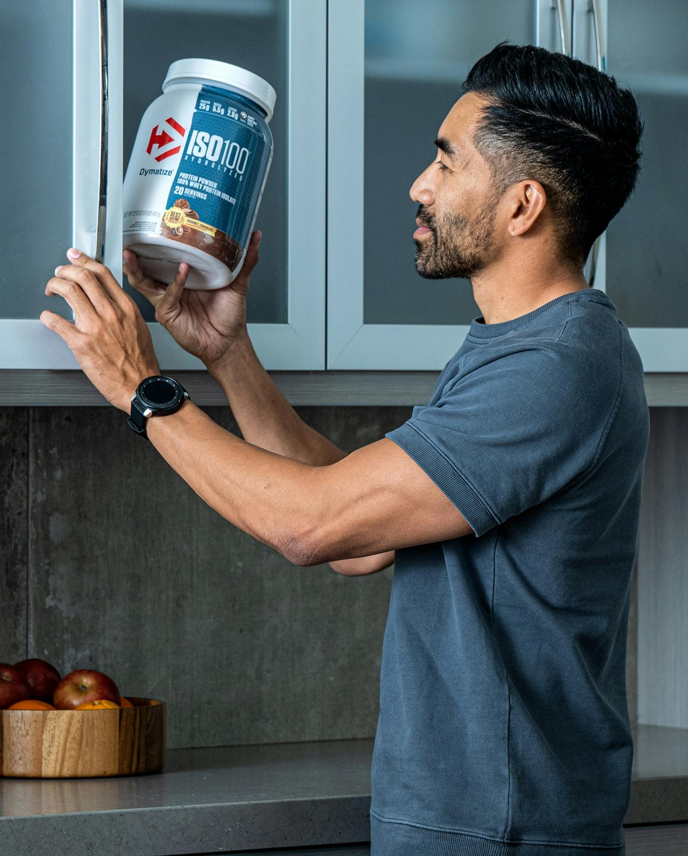 A man in his kitchen, taking a large tub of Dymatize ISO100 out of his pantry.